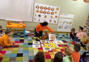 Dzieci siedzą na dywanie w półkolu, a nauczycielka prezentuje im cykl wzrostu dyni.