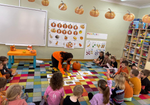 Dzieci siedzą na dywanie w półkolu, a nauczycielka zapisuje na kartce ich skojarzenia z dynią.