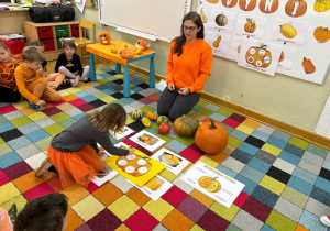 Dzieci siedzą na dywanie w półkolu, a dziewczynka przykleja karteczkę z jednym etapów cyklu rozwoju dyni.