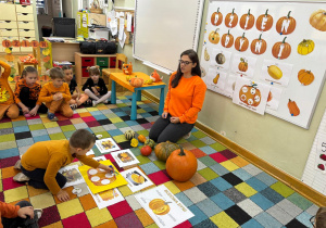 Dzieci siedzą na dywanie w półkolu, a chłopiec przykleja kolejną karteczkę z jednym z etapów cyklu rozwoju dyni.