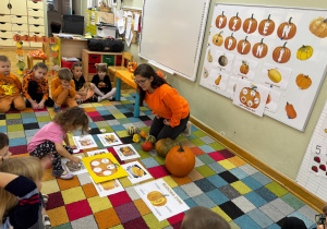 Dzieci siedzą na dywanie w półkolu, a dziewczynka przykleja następną karteczkę z jednym z etapów cyklu rozwoju dyni.