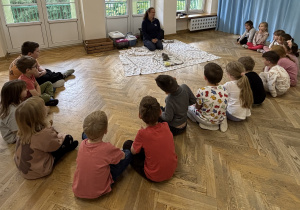 Dzieci siedzą w półkolu i słuchają, jak Pani opowiada im o prezentowanym zwierzątku tj. kurce.