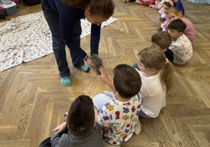 Dzieci siedzą w półkolu i głaszczą kurkę, którą trzyma w rękach Pani prowadząca zajęcia.