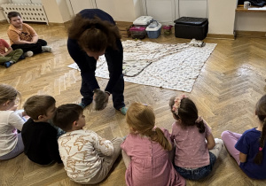 Dzieci siedzą w półkolu i głaszczą kurkę, którą trzyma w rękach Pani prowadząca zajęcia.