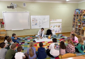 Dzieci siedzą na dywanie w półkolu, a prowadząca gra na kieliszku z wodą.