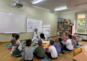 Dzieci siedzą na dywanie w półkolu, a prowadząca trzyma rękę na swojej szyi i prezentuje dzieciom, że dźwięk to fale, które powodują drgania.