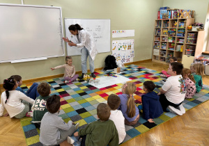 Dzieci siedzą w półkolu, prowadząca klaszcze nad głową jednej dziewczynki, która siedzi po środku z zamkniętymi oczami i musi zgadnąć skąd dobiega klaśnięcie.