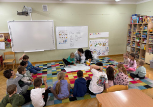 Dzieci siedzą na dywanie w półkolu, a prowadząca z jednym chłopcem grają na kieliszku z wodą.