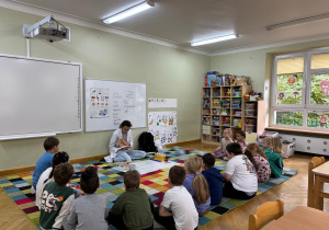 Dzieci siedzą w półkolu na dywanie, a prowadząca warsztaty prezentuje im model ludzkiego ucha i nazywa jego elementy.