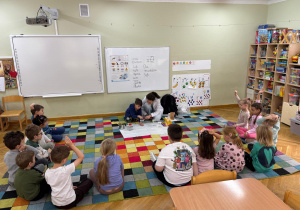 Dzieci siedzą na dywanie w półkolu, a prowadząca z jednym chłopcem grają na kieliszku z wodą.