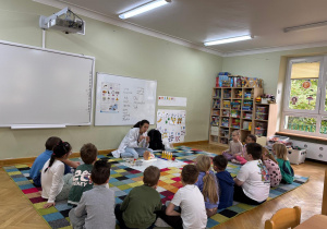 Dzieci siedzą w półkolu na dywanie, a prowadząca warsztaty prezentuje im model ludzkiego ucha i nazywa jego elementy.