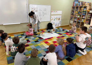Dzieci siedzą w półkolu, prowadząca klaszcze nad głową jednej dziewczynki, która siedzi po środku z zamkniętymi oczami i musi zgadnąć skąd dobiega klaśnięcie.