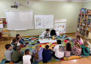 Dzieci siedzą w półkolu, a prowadząca prezentuje doświadczenie, jak rozchodzi się dźwięk, stukając drewnianą łyżką w garnek.