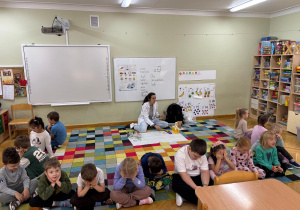 Dzieci siedzą w półkolu na dywanie odwrócone plecami do prowadzącej i próbują odgadnąć na jakim instrumencie Pani gra przy użyciu samego słuchu.