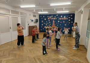 Dzieci stoją ustawione w rozsypce na sali gimnastycznej i powtarzają kroki taneczne za prowadzącą zajęcia z zumby.