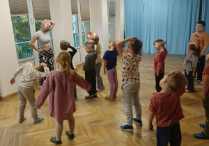 Dzieci stoją w rozsypce z jedną ręką na głowie, a drugą na boku i wykonują ćwiczenia rozciągające na zajęciach zumby.