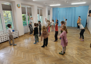 Dzieci stoją w rozsypce z jedną ręką uniesioną do góry pochylają się do boku.
