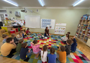 Dzieci siedzą na dywanie w kole, a nauczycielka pokazuje im swoje ręce i pyta, czy bakterie są widoczne.