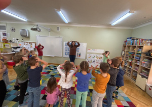 Dzieci stoją w kole na dywanie i powtarzają za nauczycielką ćwiczenia gimnastyczne na rozgrzewkę.