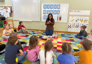 Dzieci siedzą w kole na dywanie i obserwują, jak nauczycielka pokazuje pieprz, który będzie "bakteriami".
