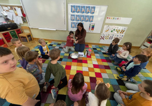 Dzieci siedzą w kole na dywanie i obserwują, jak dziewczynka zanurza palec w mydle i wykonuje doświadczenie, jak "bakterie" uciekają od mydła.