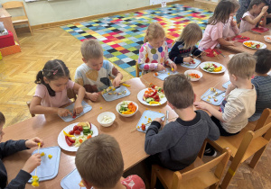 Dzieci siedzą przy stolikach i wykonują samodzielnie szaszłyki z warzyw podczas Dnia Owoców i Warzyw.