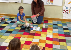 Dzieci siedzą w kole na dywanie i obserwują jak dziewczynka myje rączki pocierając rączki o siebie rozprowadzając mydło po nich.