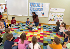 Dzieci siedzą w kole na dywanie i obserwują jak dziewczynka myje rączki spłukując mydło wodą.