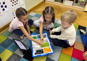 Dwie dziewczynki i chłopiec siedzą na dywanie i budują z klocków Lego na zajęciach z robotyki.