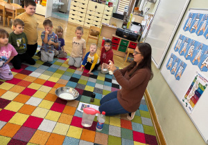 Dzieci siedzą w kole na dywanie i obserwują, jak nauczycielka nalewa mydło do szklanki.