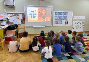 Dzieci siedzą na dywanie i oglądają filmik edukacji nt. mycia rąk.