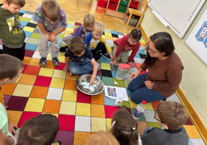 Dzieci siedzą w kole na dywanie i obserwują, jak chłopiec wykonuje doświadczenie, jak "bakterie" uciekają od mydła.