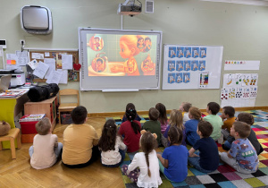 Dzieci siedzą na dywanie i oglądają filmik edukacji nt. mycia rąk.