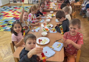 Dzieci siedzą przy stolikach i prezentują wykonane przez siebie szaszłyki z warzyw podczas Dnia Owoców i Warzyw.