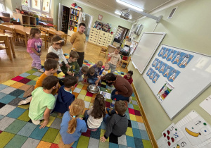 Dzieci siedzą w kole na dywanie i obserwują, jak chłopiec wykonuje doświadczenie, jak "bakterie" uciekają od mydła.