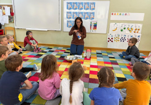 Dzieci siedzą w kole na dywanie i obserwują, jak nauczycielka pokazuje mleko, które będzie wykorzystane do doświadczenia..