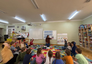 Dzieci siedzą na dywanie w kole, nauczycielka zadaje im pytania, a dzieci podnoszą ręce do góry chcąc odpowiedzieć na nie.