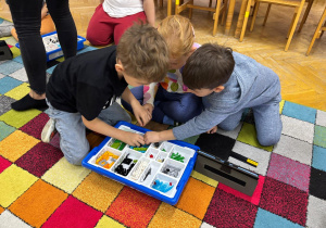 Dwóch chłopców i dziewczynka siedzą na dywanie i budują z klocków Lego na zajęciach z robotyki.