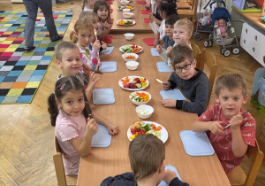 Dzieci siedzą przy stolikach trzymają noże w dłoni i przygotowują się do robienia szaszłyków.