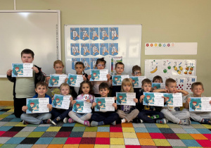 Dzieci siedzą na dywanie i prezentują swoje dyplomy za aktywny udział w zajęciach z okazji Światowego Dnia Mycia Rąk.