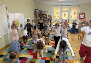 Dzieci stoją na sali w rozsypce i podskakują do góry i robią skłon do przodu przy muzyce.