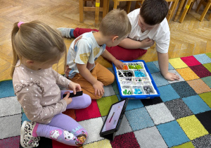 Dziewczynka i chłopcy siedzą na dywanie i budują z klocków Lego na zajęciach z robotyki.