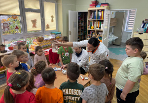 Dzieci stoją przy stoliku i obserwują, jak mama kolegi wykonuje eksperyment, który pokazuje, jak rozpuszcza się tabletka w naszym żołądku.