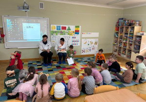 Dzieci siedzą na dywanie w półkolu i rozmawiają z rodzicami kolegi, którzy są farmaceutami nt. pracy w aptece podczas wizyty rodziców kolegi w ramach projektu Bohater Tygodnia.