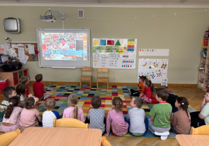 Dzieci siedzą na dywanie w półkolu i oglądają filmik edukacyjny nt. pracy w aptece w ramach projektu Bohater Tygodnia.