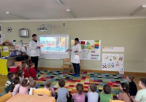 Dzieci siedzą na dywanie w półkolu, a rodzice kolegi prezentują przygotowany filmik edukacyjny nt. pracy w aptece w ramach projektu Bohater Tygodnia.