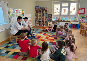 Dzieci siedzą na dywanie w półkolu i rozmawiają z rodzicami kolegi, którzy są farmaceutami nt. rodzajów dostępnych leków podczas wizyty rodziców kolegi w ramach projektu Bohater Tygodnia.