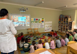 Dzieci siedzą na dywanie w półkolu i oglądają filmik edukacyjny nt. pracy w aptece w ramach projektu Bohater Tygodnia.