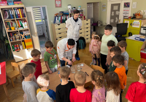 Dzieci stoją przy stoliku i obserwują, jak mama kolegi wykonuje eksperyment, który pokazuje, jak rozpuszcza się tabletka w naszym żołądku.