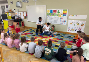 Dzieci siedzą na dywanie w półkolu, a kolega wspólnie z rodzicami prezentuje przygotowaną książeczkę w ramach projektu Bohater Tygodnia.